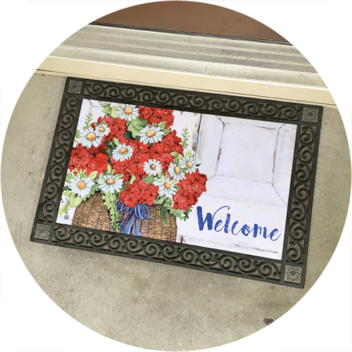 A door mat with red and white flowers and blue writing saying Welcome.